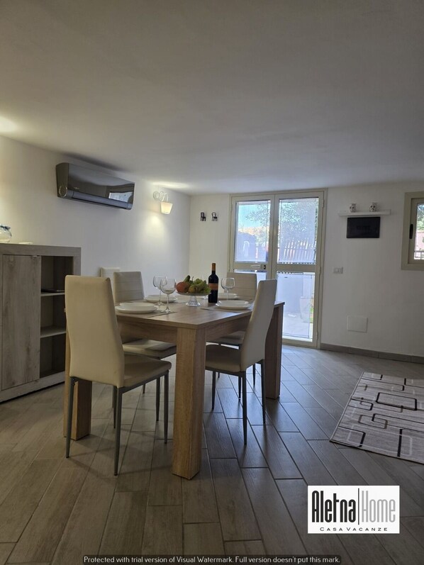 A dining area with a wooden table set for four, surrounded by chairs. There is a buffet table with fruits and a bottle of wine. The room has a large window, a wall-mounted air conditioner, and a small shelf with decorative items. - ALETNA HOME vacation home (Ragalna)