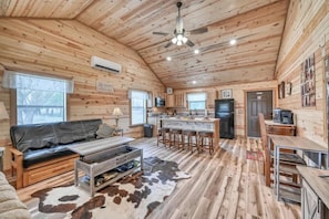 A cabin interior with a wooden ceiling, a black leather sofa, a kitchen area, and a dining table. - Opal Cabin-HotTub-10 min to Main Street! (Fredericksburg)