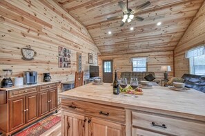 A wooden cabin interior with a kitchen area, wooden cabinets, a dining table with chairs, and a ceiling fan. - Opal Cabin-HotTub-10 min to Main Street! (Fredericksburg)