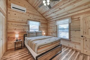 A wooden cabin bedroom with a bed, two windows, an air conditioning unit, and a ceiling fan.
