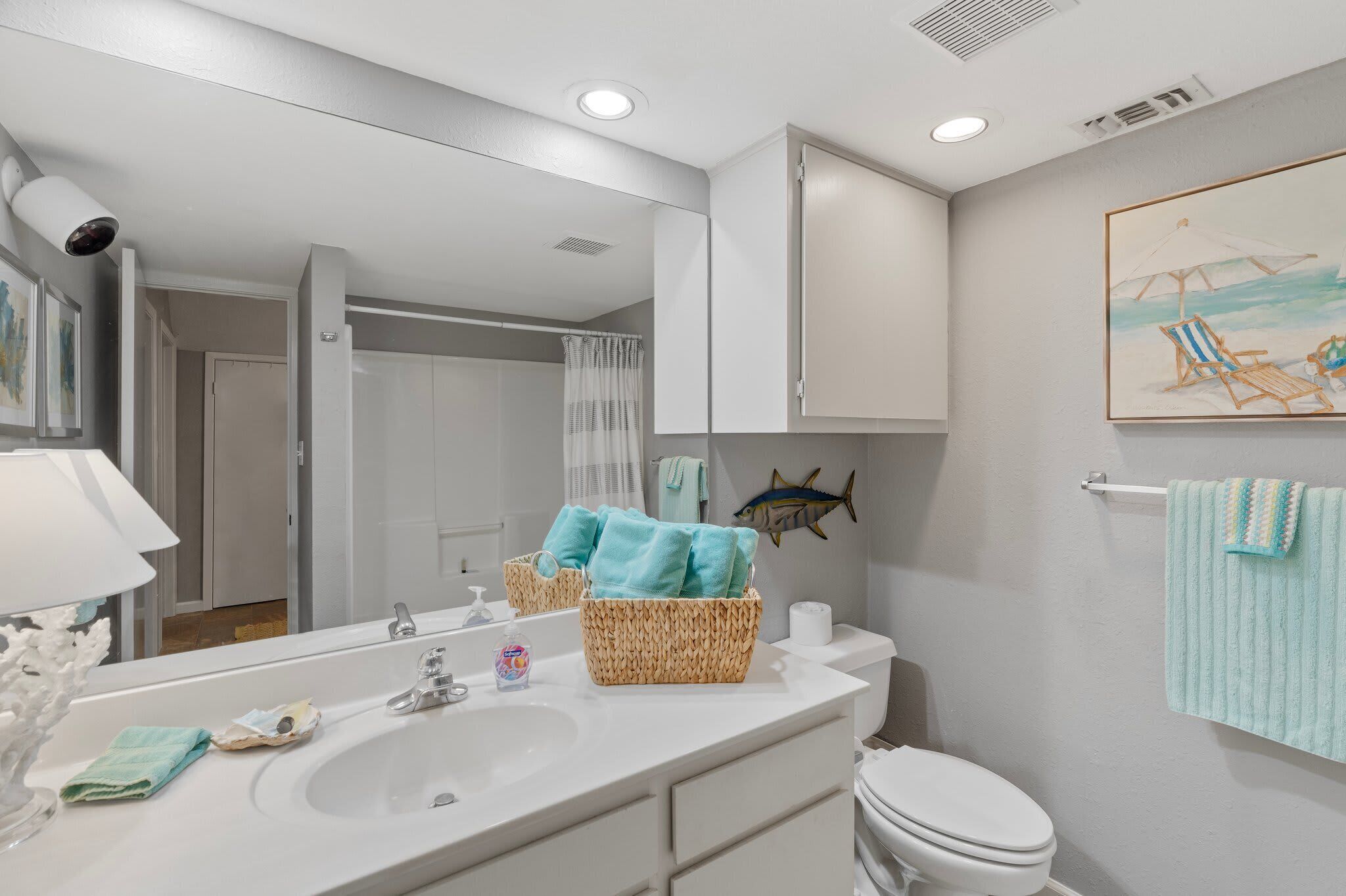 A bathroom with a white sink, a mirror, a basket of towels, a fish decoration, and a beach-themed painting.