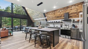 A modern kitchen with a central island, bar stools, and a large window overlooking a deck. - Luxury A-Frame*Hot-tub*Firepit (Omaha)