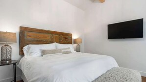 Un dormitorio con cabecera de madera, un televisor de pantalla plana en la pared, una mesita redonda con una lámpara y una cama grande con ropa de cama blanca.