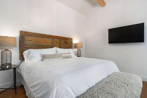 Une chambre à coucher avec une tête de lit en bois, un téléviseur à écran plat fixé au mur, une petite table ronde avec une lampe, et un grand lit avec une literie blanche.