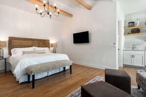A bedroom with a wooden headboard, a bed with white bedding, a bench, a chandelier, a TV on the wall, and a kitchenette with a shelf. - The Stables Suite Two-1 block to Main Street! (Fredericksburg)