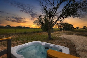 A hot tub with a wooden deck, surrounded by a rural landscape with fields and trees.