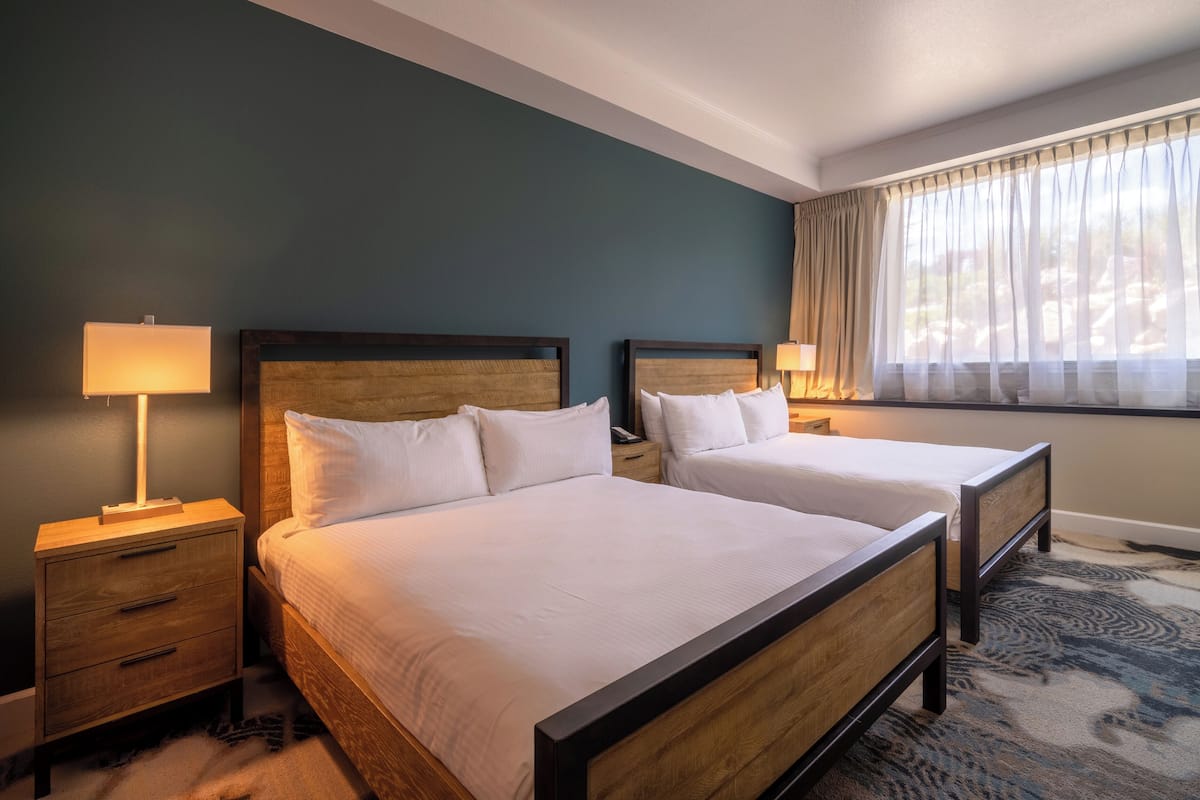 A hotel room with two beds, a nightstand with a lamp, and a window with curtains.