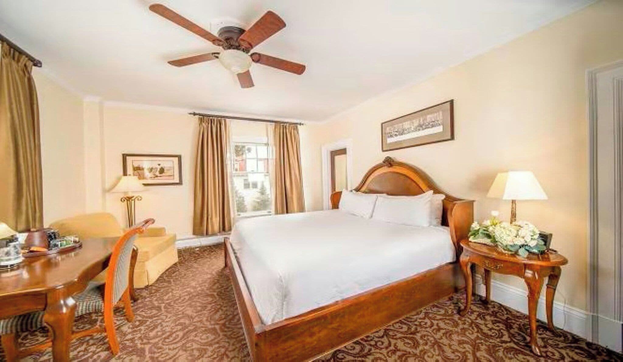 A hotel room with a bed, a desk, a chair, a ceiling fan, and a window with curtains.