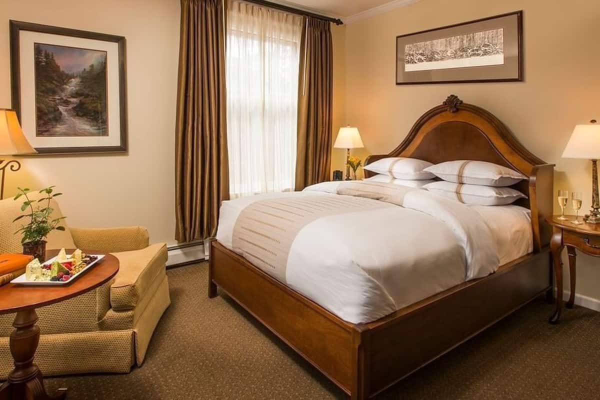 A hotel room with a large bed, two bedside tables with lamps, a chair, a small table with a tray of snacks, and two framed pictures on the walls.