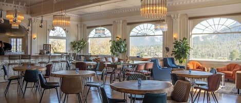 A spacious dining area with round tables and chairs, large windows with arched tops, and hanging chandeliers.