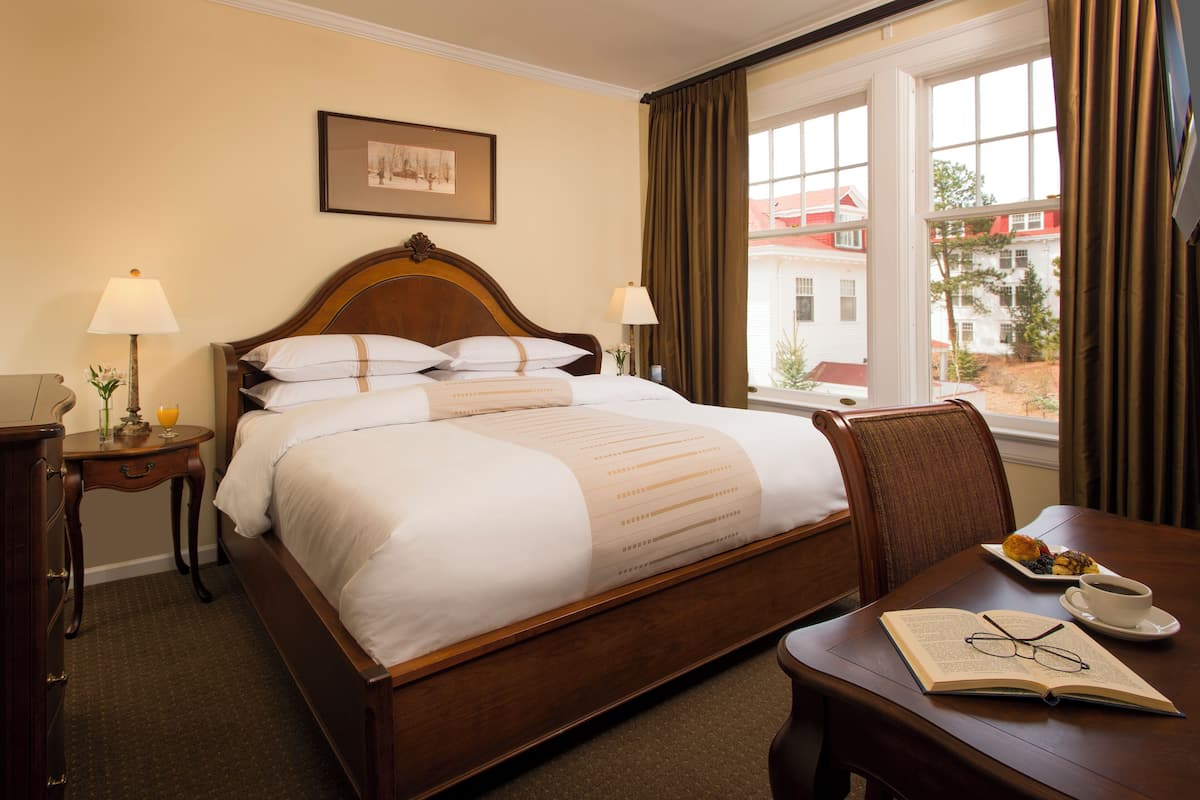A hotel room with a bed, bedside lamps, a dining table with a book and glasses, and a window with curtains.