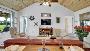 A spacious living room with a high ceiling, a large window, a ceiling fan, a fireplace, and a dining area with a table set for two.