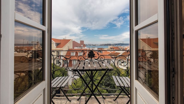 Un balcon doté d’une balustrade en fer forgé et d’une table en bois, offrant une vue sur un paysage urbain avec des bâtiments aux toits rouges.