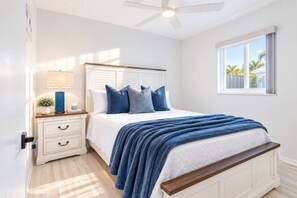 Une chambre à coucher avec un lit, une table de chevet, une lampe et une fenêtre donnant sur des palmiers.