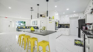 A modern kitchen with white cabinets, a central island with yellow stools, and a dining area with a sofa and coffee table. - Golden Vibes • Fun for Everyone (Bradenton)