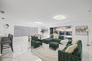 Un salon moderne avec un canapé d’angle, un coin repas comprenant une table et des chaises, et une grande fenêtre avec des stores.