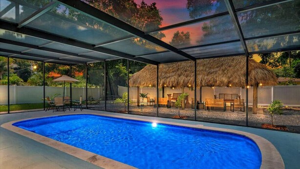 A swimming pool with a glass enclosure, a thatched-roof structure, and outdoor seating. - Golden Vibes • Fun for Everyone (Bradenton)