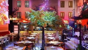 An indoor dining area with tables set for meals, a central decorative tree, and a background with a mural and windows. - Condo in Epic Punta Cana! Near Cocobongo (Punta Cana)