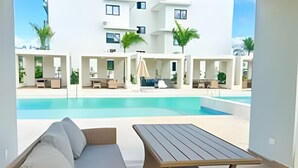 A modern outdoor seating area with a pool, white buildings in the background, and a clear sky. - Condo in Epic Punta Cana! Near Cocobongo (Punta Cana)