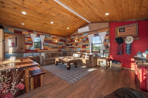 A cozy living room with a wooden ceiling, a colorful wall, and a red accent wall with an American flag. There are two sofas, a coffee table, and a dining table with benches.