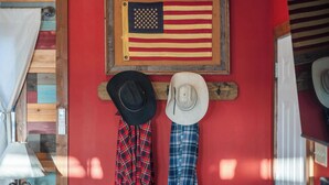 Una habitación con pared roja, puerta de madera y una bandera estadounidense colgada. Hay dos sombreros y dos camisas de cuadros colgados en la pared.