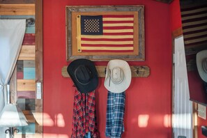 A room with a red wall, a wooden door, and a mounted American flag. There are two hats and two plaid shirts hanging on the wall.