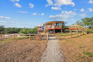 A rustic cabin with a wooden deck and a red roof, surrounded by a gravel path and a wooden fence.