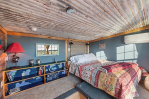 A bedroom with a rustic ceiling, a bed with a colorful quilt, a wooden bench, a shelf with books, and a window with a view.