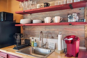 A kitchen with a stainless steel sink, a red coffee maker, and shelves with cups and plates.
