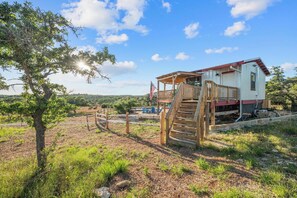 A rustic cabin with a wooden deck and a small porch, surrounded by a fenced yard and a tree.