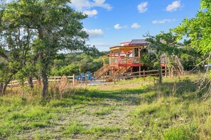 A wooden cabin with a balcony and a flag, surrounded by trees and grass. - 20-Acre Ranch Getaway: Heated Spool, Firepit, (Blanco)