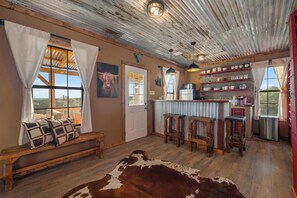 Un intérieur rustique avec un banc en bois, un tapis en peau de vache et un coin cuisine doté d’étagères et de tabourets.
