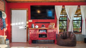 Una sala de estar con una pared roja, una puerta blanca, un televisor de pantalla plana, un mueble de madera para equipo audiovisual, un sillón tipo puff y una ventana con cortinas blancas.