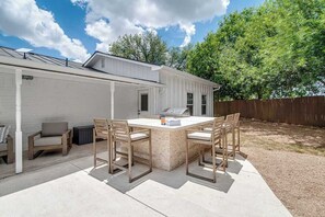 Un jardin avec une terrasse en béton, une table en bois et quatre chaises, un barbecue et une maison blanche dotée d’un porche abrité.