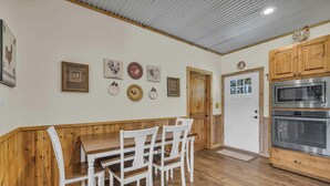 Una cocina con armarios de madera, un microondas integrado y una zona de comedor con mesa de madera y sillas blancas.