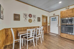 Una cocina con armarios de madera, un microondas integrado y una zona de comedor con mesa de madera y sillas blancas.