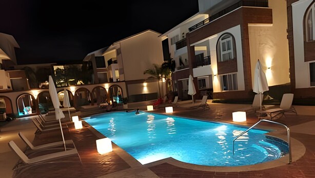 Ein Pool mit versenktem Zentrum, umgeben von Liegestühlen und Sonnenschirmen, mit Gebäuden im Hintergrund.