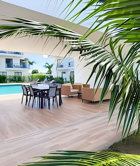 A modern outdoor dining area with a pool, wooden flooring, and palm trees.