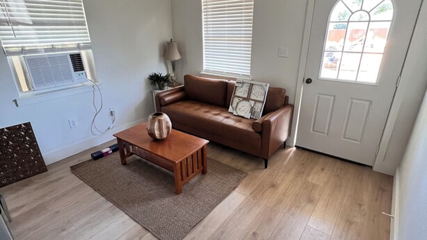 A spacious living room with a brown leather sofa, a wooden coffee table, and a chandelier. There are two windows with blinds, a door with a glass panel, and a white wall. - Cozy cabin in nice Wylie with AC (Wylie)