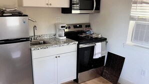 A compact kitchen with white cabinets, a stainless steel refrigerator, a microwave, an oven, and a coffee maker. There's a wooden table with a vase and a rug on the floor. - Cozy cabin in nice Wylie with AC (Wylie)