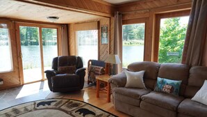 Un interior de cabaña acogedor, con un sillón de cuero, un sofá con cojines decorativos, una mesa de madera y ventanas amplias que dan a un lago.