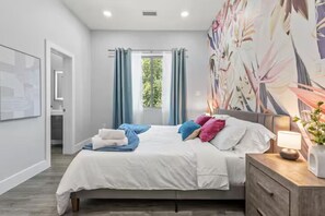 A modern bedroom with a bed, a nightstand, a wall-mounted lamp, and a window with curtains. - Sleeps 12! Hot Tub & Game Room Near Downtown! (Dallas)