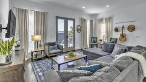 A modern living room with a grey sofa, a coffee table, and a TV on a stand. There are curtains on the windows, a plant on the side table, and a guitar on the wall.