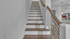 A staircase with wooden steps and a wooden handrail, leading to a room with a white wall and a modern kitchen area.