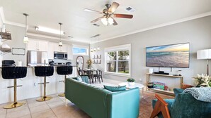 A modern kitchen with white cabinets, a dining area with a green sofa, and a living room with a TV and a bookshelf.