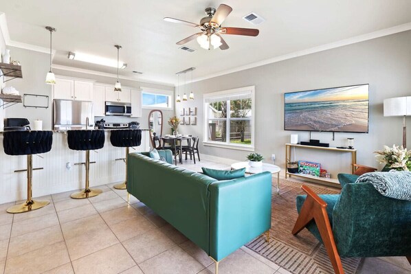A modern kitchen with white cabinets, a dining area with a green sofa, and a living room with a TV and a bookshelf.