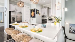 A modern kitchen with white cabinetry, stainless steel appliances, a central island with bar stools, and decorative wall art. - 5 Mins from Beach with Private Golf Cart and Pool (Santa Rosa Beach)