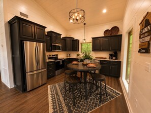 A modern kitchen with dark cabinetry, stainless steel appliances, and a central island with stools. - 1BR Cabin • HotTub & Fire Pit |BBQ | Patio | Pets (Broken Bow)