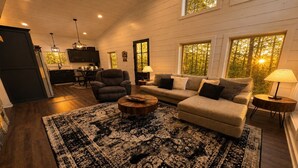 A cozy living room with a sofa, armchair, and coffee table, wooden floors, and large windows with a view of trees. - Romantic Cabin with Hot Tub, Fire Pit, BBQ & Fenced Yard (Broken Bow)