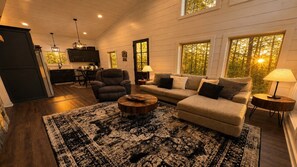 A cozy living room with a sofa, armchair, and coffee table, wooden floors, and large windows with a view of trees. - 1BR Cabin | HotTub | Fire Pit | BBQ | Patio | Pets (Broken Bow)